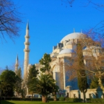 Mavi gökyüzü altında, ağaçların arkasında yükselen kubbeli ve minareli Süleymaniye Camii