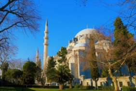 Mavi gökyüzü altında, ağaçların arkasında yükselen kubbeli ve minareli Süleymaniye Camii