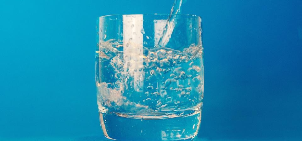 Mavi bir fonun önünde, içine su dökülürken sıçrayan damlalarla dolu şeffaf bir su bardağı